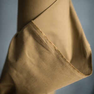 May include: A roll of tan fabric, with a visible edge showing the stitching. The fabric is tightly rolled, with a portion of the material unfurled, revealing its texture and color. The background is a muted blue.