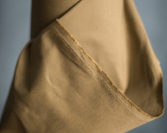 May include: A roll of tan fabric, with a visible edge showing the stitching. The fabric is tightly rolled, with a portion of the material unfurled, revealing its texture and color. The background is a muted blue.