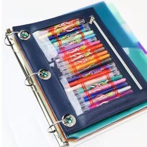 May include: A dark blue three-ring binder featuring a clear plastic front pocket filled with a collection of colorful pens. The binder has silver metal rings and a zippered closure along the side of the transparent storage compartment.