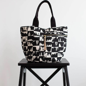 May include: Black and white tote bag with a brushstroke pattern. The bag has black handles and a zippered pocket with a gold zipper and a brown tassel.