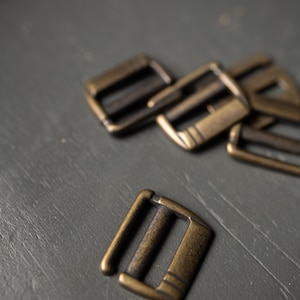 May include: Close-up of several antique brass-colored metal slider buckles. The buckles have a rectangular shape with a central bar and decorative lines. The background is a muted gray-green surface.