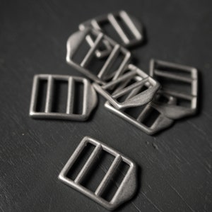 May include: A close-up of a pile of silver metal slider buckles. The buckles are rectangular with three horizontal bars and a rounded edge.