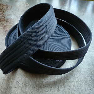 May include: A roll of black webbing, a type of heavy-duty fabric tape, is coiled up on a table. The webbing is woven with a textured pattern and appears to be strong and durable.