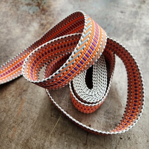 May include: A coiled roll of colorful woven ribbon. The ribbon features a repeating pattern of orange, purple, blue, and white geometric shapes. The edges are white, and the ribbon is displayed on a textured, metallic surface.