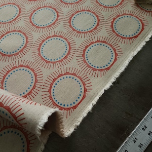 May include: A beige fabric featuring a repeating pattern of red sunburst motifs with blue dotted circles in the center. A metal ruler is visible along the edge, showing measurements in inches, with the fabric width spanning approximately 22 inches.
