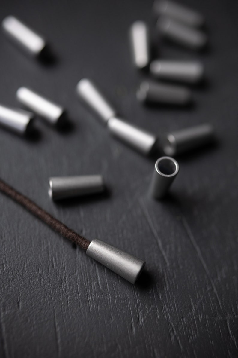 May include: A close-up of silver metal cord ends on a black surface. A brown cord is threaded through one of the cord ends.