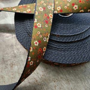 May include: A roll of dark gray webbing with a floral patterned ribbon. The ribbon features a green background with a repeating pattern of small white, red, and orange flowers. The webbing is tightly wound, and the ribbon is partially unrolled.
