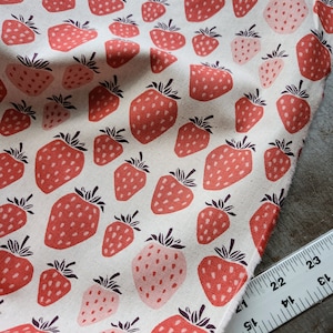 May include: A white fabric with a repeating pattern of red and pink strawberries. The fabric is folded and a ruler is visible in the bottom right corner.