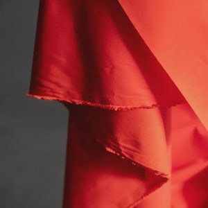 May include: Close-up of vibrant red fabric with a raw, unfinished edge. The fabric is layered, creating depth and texture. The color is a bold, saturated red, with a slight sheen. The background is a muted gray.