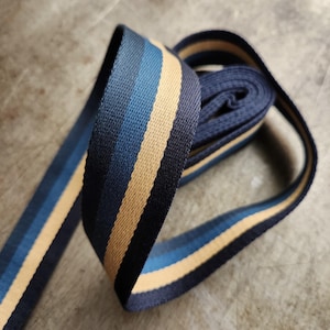 May include: Close-up of a navy blue and tan striped ribbon, partially rolled. The ribbon has a woven texture and features stripes of varying shades of blue and tan. The ribbon is on a neutral-toned surface.