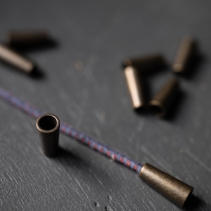 May include: A close-up of a cord with two metal cord ends. The cord is blue and red with a white stripe. The cord ends are a dark bronze color.