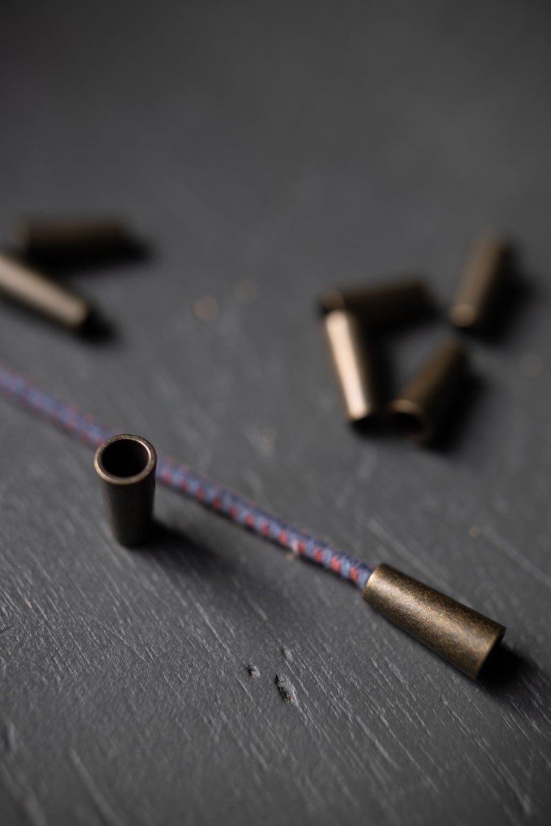 May include: A close-up of a cord with two metal cord ends. The cord is blue and red with a white stripe. The cord ends are a dark bronze color.