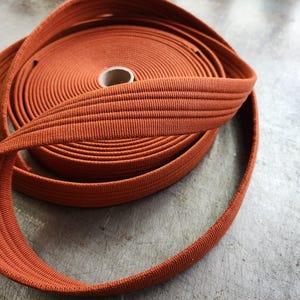 K&ouml;nnte beinhalten: Eine Rolle orangefarbenen elastischen Bandes, eng gewickelt, mit sichtbarer Textur. Das Band ist in einer Schlaufe abgewickelt, was seine Flexibilit&auml;t zeigt. Der Hintergrund ist eine strukturierte, neutrale Oberfl&auml;che.
