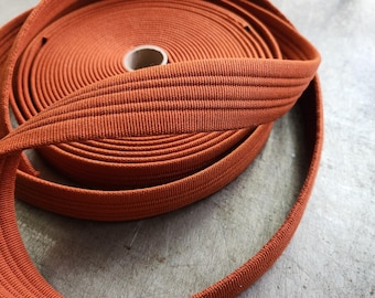 May include: A roll of orange elastic ribbon, tightly wound, with a visible texture. The ribbon is unwound in a loop, showing its flexibility. The background is a textured, neutral surface.