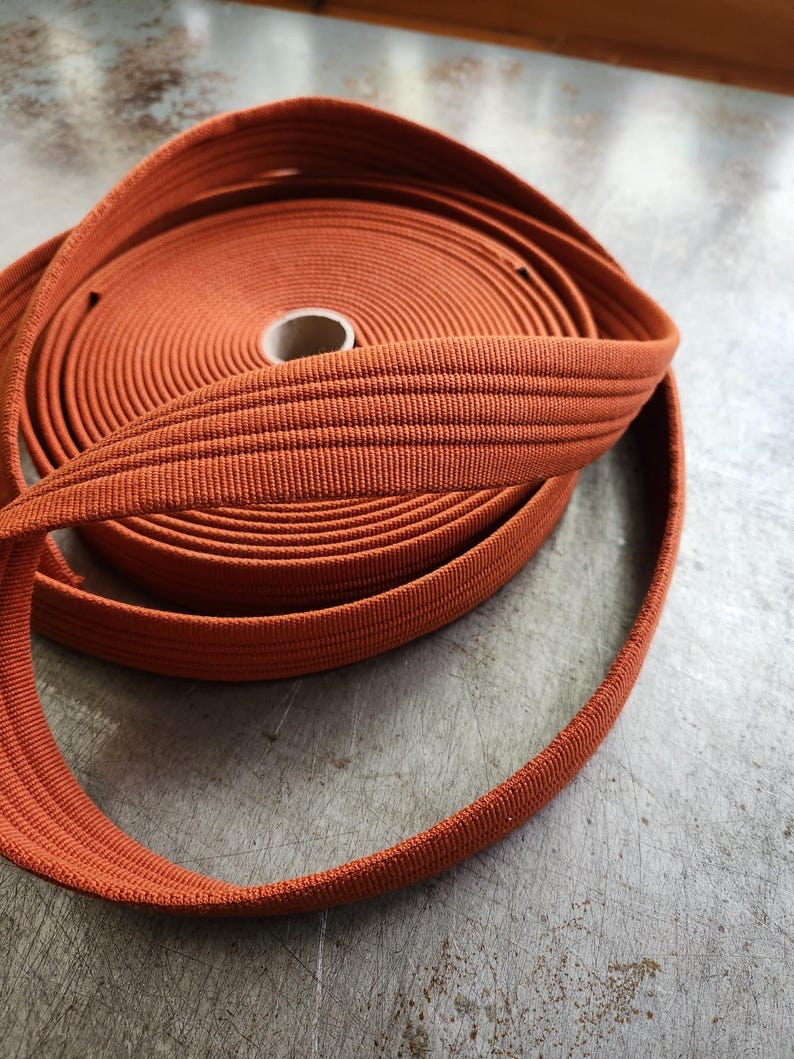 K&ouml;nnte beinhalten: Eine Rolle orangefarbenen elastischen Bandes, eng gewickelt, mit sichtbarer Textur. Das Band ist in einer Schlaufe abgewickelt, was seine Flexibilit&auml;t zeigt. Der Hintergrund ist eine strukturierte, neutrale Oberfl&auml;che.