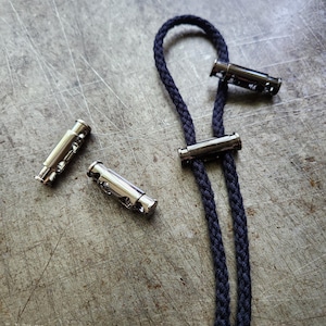 May include: A navy blue braided cord with three silver-colored cord locks. The cord is looped and has two strands. The cord locks are cylindrical with holes and are designed to secure the cord.