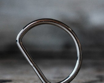 May include: A silver metal D-ring, also known as a key ring, is shown on a gray textured surface.