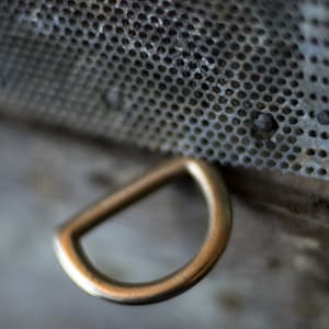 May include: A close-up of a gold-colored metal D-ring. The ring is attached to a gray metal surface with a pattern of small holes.