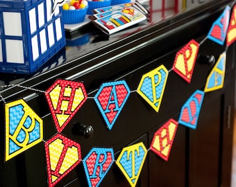 Può includere: Uno striscione colorato con le parole "Happy Birthday" scritte su bandierine a forma di diamante. Ogni bandierina ha un motivo a chevron rosso, giallo e blu con una lettera bianca al centro.