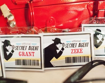 May include: Three white card badges with black and white illustrations of a person wearing a fedora and holding a magnifying glass. The text on the badges reads "Official Secret Agent" followed by the name "Grant", "Zeke", and "Chad".