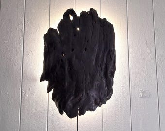 Puede incluir: Una obra de arte mural de madera oscura de forma irregular con iluminación integrada. La madera tiene una superficie texturizada con varios agujeros y una forma natural. La fuente de luz está detrás de la madera, creando un efecto brillante.