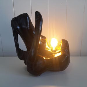 May include: A dark wood table lamp with a light bulb illuminating the inside of the lamp. The lamp is shaped like a pair of hands holding a light bulb.