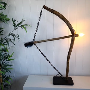 Può includere: Una lampada in legno a forma di arco e freccia. L'arco è fatto di un ramo naturale e la freccia è in legno con piume nere. La lampada ha una catena nera e una base nera.