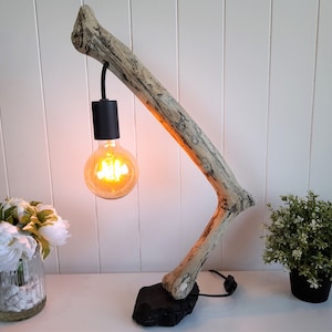 Op de afbeelding: Een rustieke tafellamp gemaakt van een natuurlijke drijfhouttak. De lamp heeft een zwarte fitting met een warm gloeiende Edison-stijl gloeilamp. De basis is een donkere, getextureerde steen, tegen een witte achtergrond.