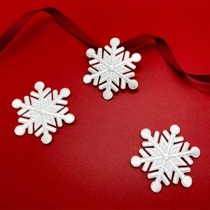 May include: Three white snowflake ornaments on a red background. The snowflakes are made of felt and have a 3D effect.