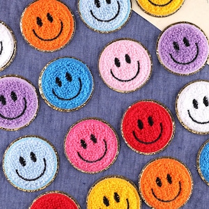 May include: A collection of circular, fuzzy patches featuring embroidered smiley faces in various colours including orange, blue, pink, red, purple, yellow, and white. Each patch has a stitched black smiling expression and a gold metallic border.