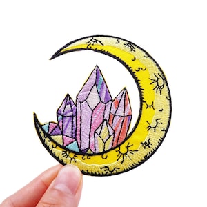 Peut inclure: Un patch brodé thermocollant représentant un croissant de lune jaune avec un groupe de cristaux violets, roses et bleus au centre. La lune est décorée d'un symbole de soleil répété.