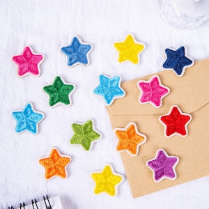 Pode incluir: Conjunto de 14 patches bordados em forma de estrela coloridos. Os patches são de várias cores, incluindo vermelho, azul, verde, amarelo e laranja. As estrelas são contornadas de branco.