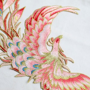 Può includere: Fenice ricamata rosa e oro con accenti blu. La fenice è in volo con le ali spiegate. Si tratta di una toppa o un'applicazione decorativa.