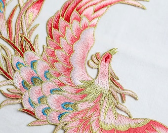 Puede incluir: Fénix bordado en rosa y dorado con detalles azules. El fénix está en vuelo con las alas extendidas. Se trata de un parche o aplique decorativo.
