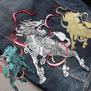 Può includere: Toppe ricamate raffiguranti creature mitiche in argento, oro e turchese. Le toppe sono cucite su una giacca di jeans, mostrando disegni dettagliati di draghi e altre bestie fantastiche con nastri fluenti.