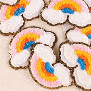 Könnte beinhalten: Ein Set mit sechs Bügelbildern in Form von Regenbögen und Wolken. Die Patches sind aus flauschigem Stoff und haben einen goldenen Glitzerbesatz. Jedes Patch zeigt einen Regenbogen in den Farben Rosa, Orange, Gelb und Blau.