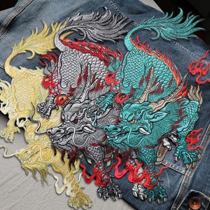 Może przedstawiać: Haftowane naszywki ze smokami w kolorze złotym, srebrnym i turkusowym z czerwonymi akcentami na dżinsowej kurtce. Szczegółowe naszywki przedstawiają mityczne stworzenia ze skomplikowanymi wzorami, pazurami i płynącymi grzywami. Naszywki są przyszyte do niebieskiej kurtki dżinsowej.