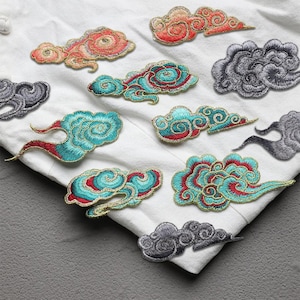 Pode incluir: Aplicações de nuvens bordadas em várias cores, incluindo turquesa, vermelho, dourado e cinza, dispostas sobre um tecido branco. As aplicações apresentam costuras detalhadas e desenhos em espiral, adequadas para decoração de tecidos ou projetos de artesanato.