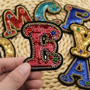 May include: A red letter E patch featuring red sequins, gold beaded trim, and small blue and green circular accents. The patch has a black border and is held in place, with other colourful sequined letter patches visible in the background.