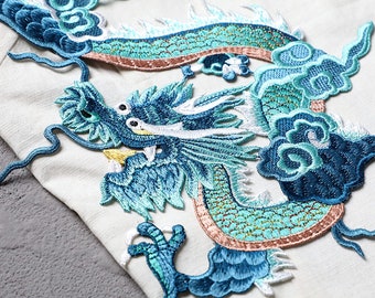 May include: A blue and green embroidered dragon design with a white background. The dragon is curled up with its head facing the left. The design is detailed and intricate.