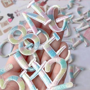 May include: Embroidered pastel-colored numbers and letters in various sizes. The patches feature a rainbow of colors, including pink, blue, yellow, and green. The patches are made of a soft, textured material.