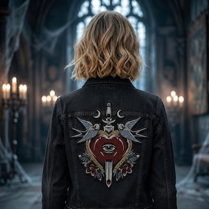 May include: A black denim jacket with a large embroidered patch on the back. The patch features a red heart with an eye, a dagger, two blue swallows, and floral accents. The jacket is worn by a person with blonde hair.
