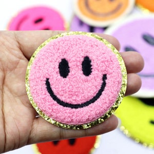 May include: A circular pink patch with a textured, fuzzy surface featuring a black embroidered smiley face. The patch is bordered by a thin, reflective gold metallic trim. Other similar circular patches in various colors are visible in the background.