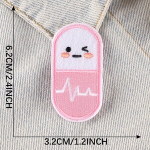 May include: Pink and white embroidered iron-on patch shaped like a pill with a heartbeat graphic and a cartoon face. The patch is 1.2 inches wide and 2.4 inches tall.