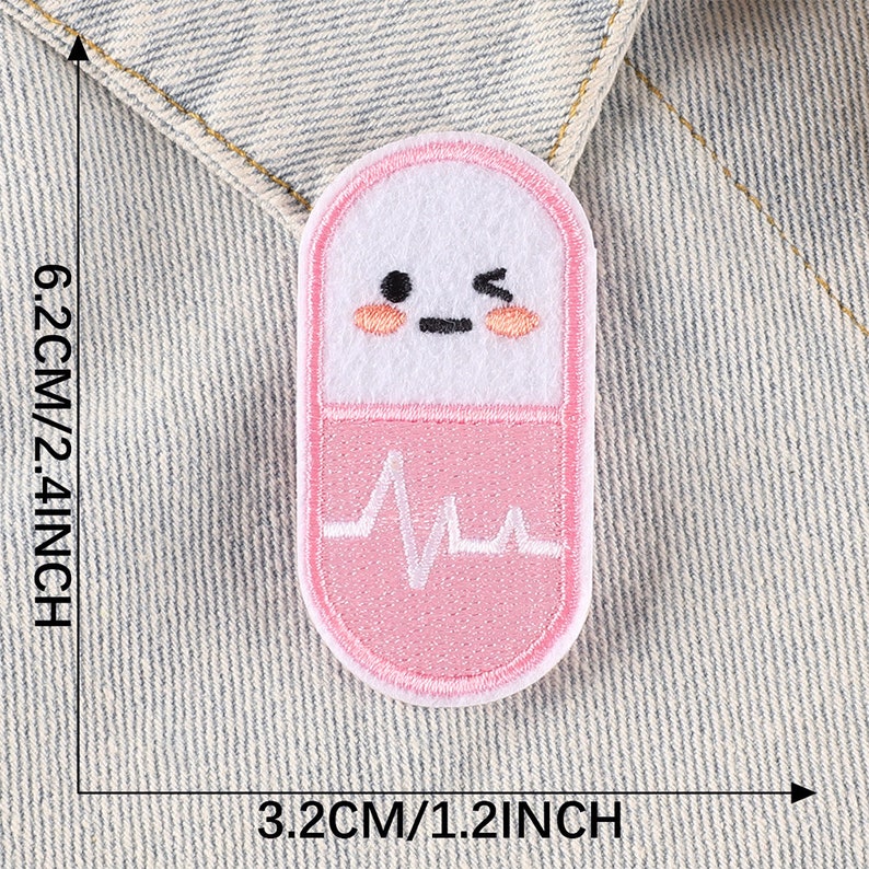 May include: Pink and white embroidered iron-on patch shaped like a pill with a heartbeat graphic and a cartoon face. The patch is 1.2 inches wide and 2.4 inches tall.