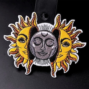 May include: Embroidered patch depicting a sun and moon with human faces. The sun is yellow with orange rays and the moon is grey with a crescent moon shape on its forehead. Both faces have closed eyes.
