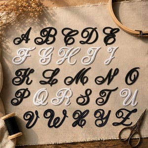 May include: A collection of embroidered alphabet letters from A to Z in a cursive script. The letters alternate between black and white thread colors, arranged in rows on a light beige fabric background, accompanied by a spool of black thread and scissors.
