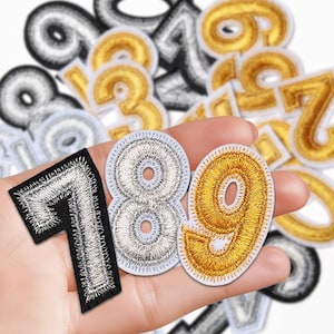 Peut inclure: Écussons de chiffres brodés en or, argent et noir. Les chiffres 7, 8 et 9 sont mis au point, les autres sont flous. Ces écussons sont destinés aux travaux manuels ou aux projets de personnalisation.