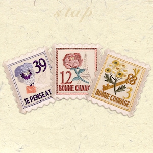 Op de afbeelding: Drie geborduurde stoffen patches met een vintage postzegelontwerp. De eerste patch heeft een paarse viooltje, een witte envelop en de tekst "Je Penseat 39". De tweede patch heeft een roze roos, de Eiffeltoren en de tekst "12 Bonne Chang". De derde patch heeft een gele bloem, een sleutel en de tekst "Bonne Courage 3".