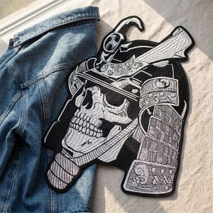 Könnte beinhalten: Ein schwarz-weißer, gestickter Aufnäher mit einem Totenkopf, der einen Samurai-Helm trägt. Der Aufnäher weist detaillierte Stickereien auf, wobei die Zähne des Schädels und das komplizierte Design des Helms deutlich zu sehen sind. Der Aufnäher liegt auf einer Jeansjacke.
